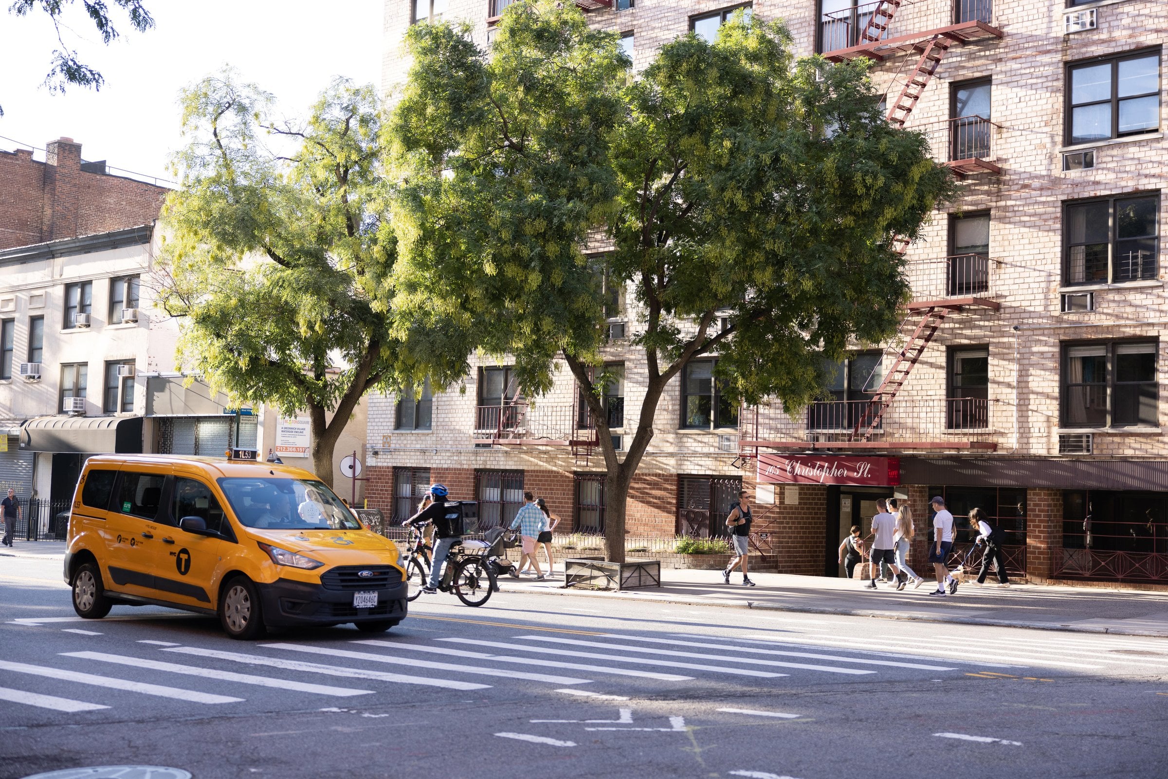 Yellow taxi and pedestrians beneath trees beside a brick building
