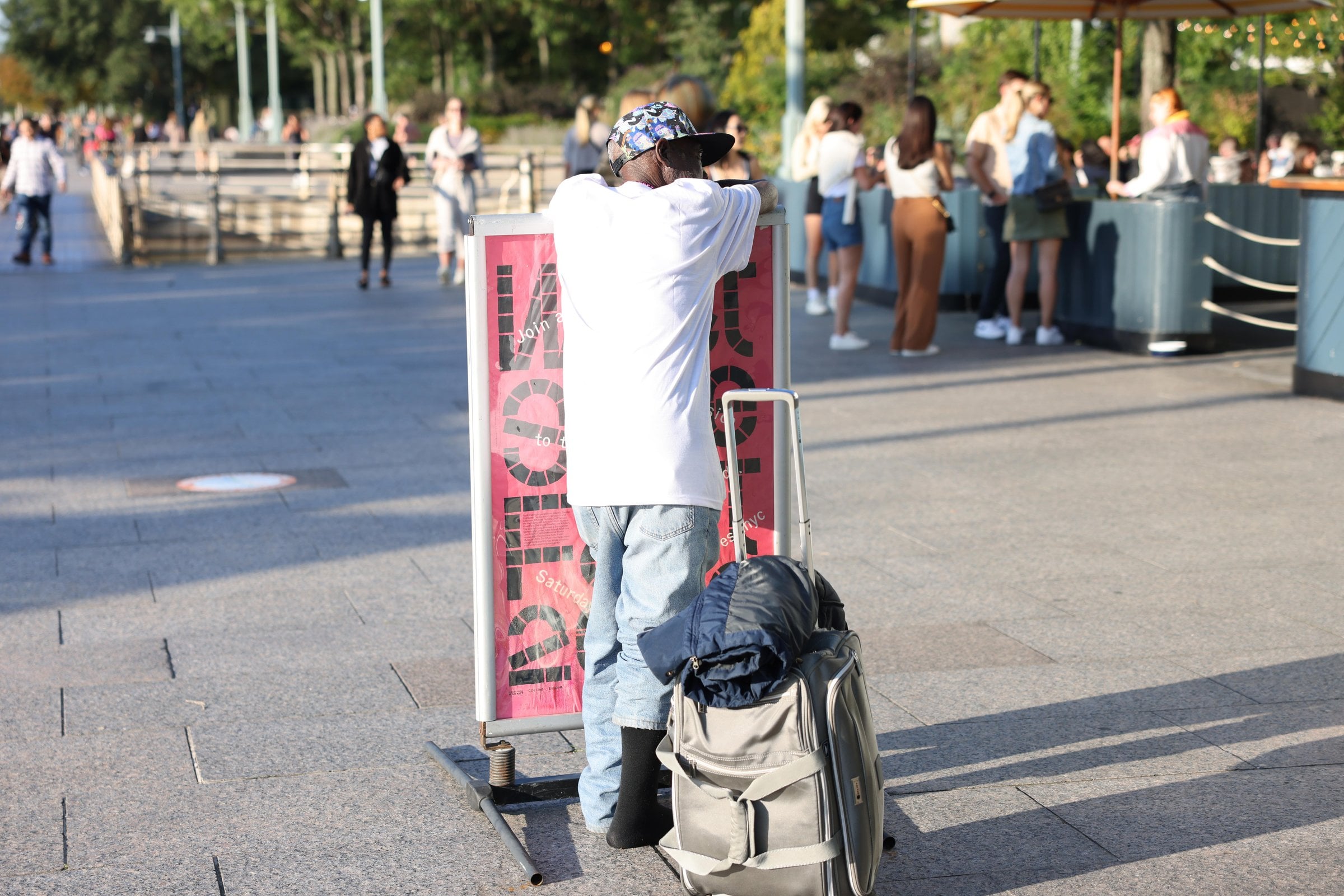 Person leaning on a sign beside a suitcase on a promenade