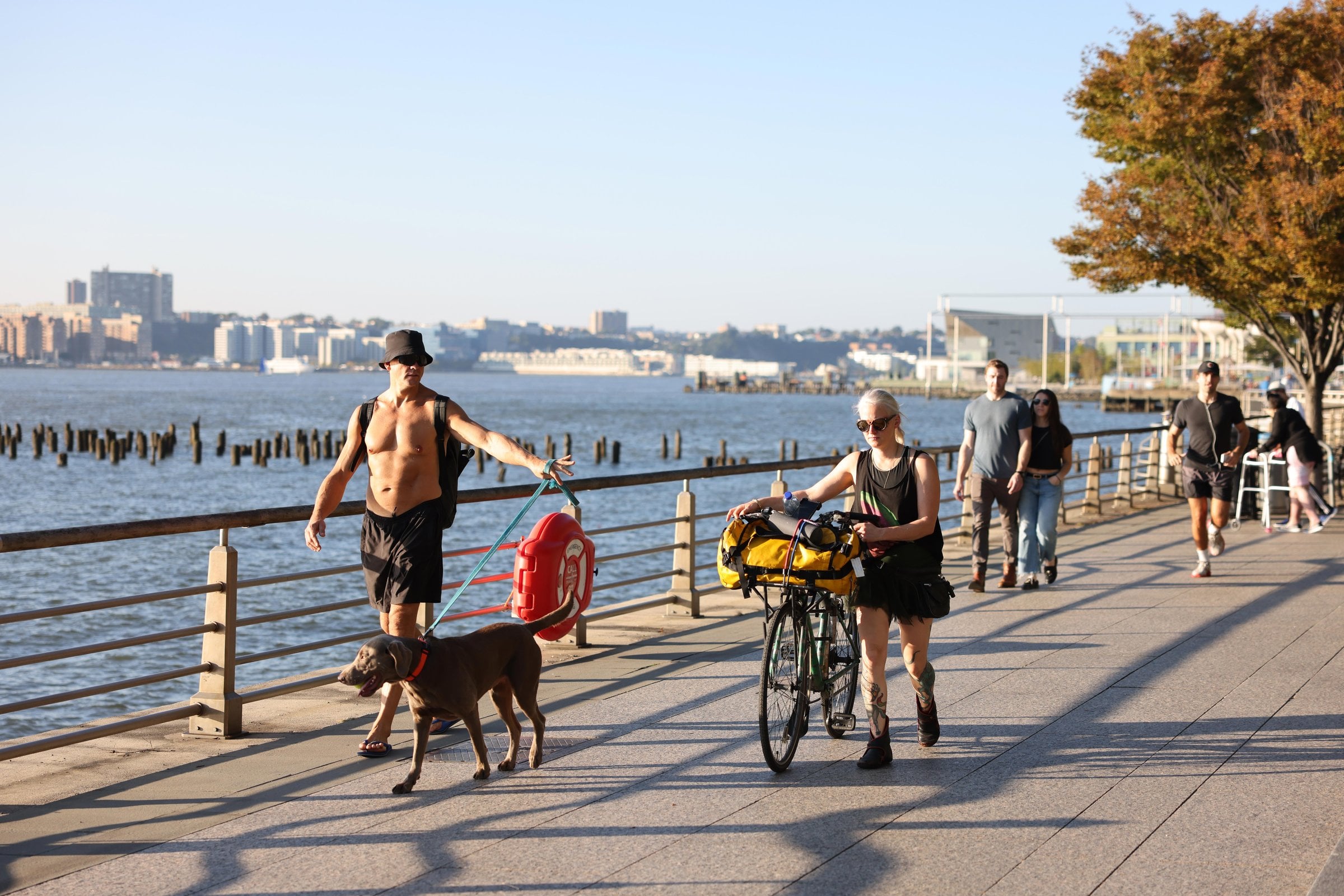 People walking with a dog and bicycle along a sunlit waterfront