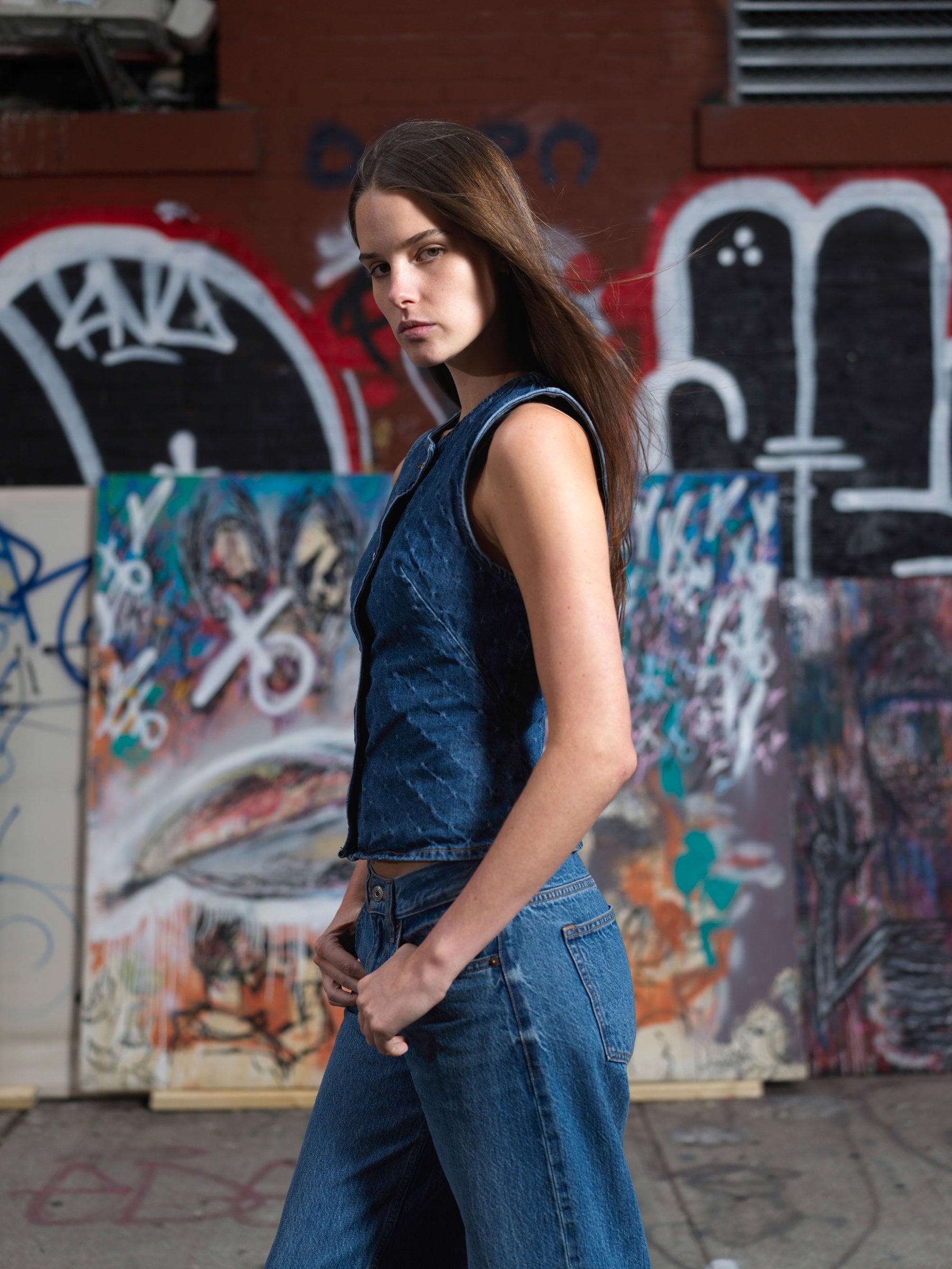 Person wearing a denim top and jeans before a graffiti wall