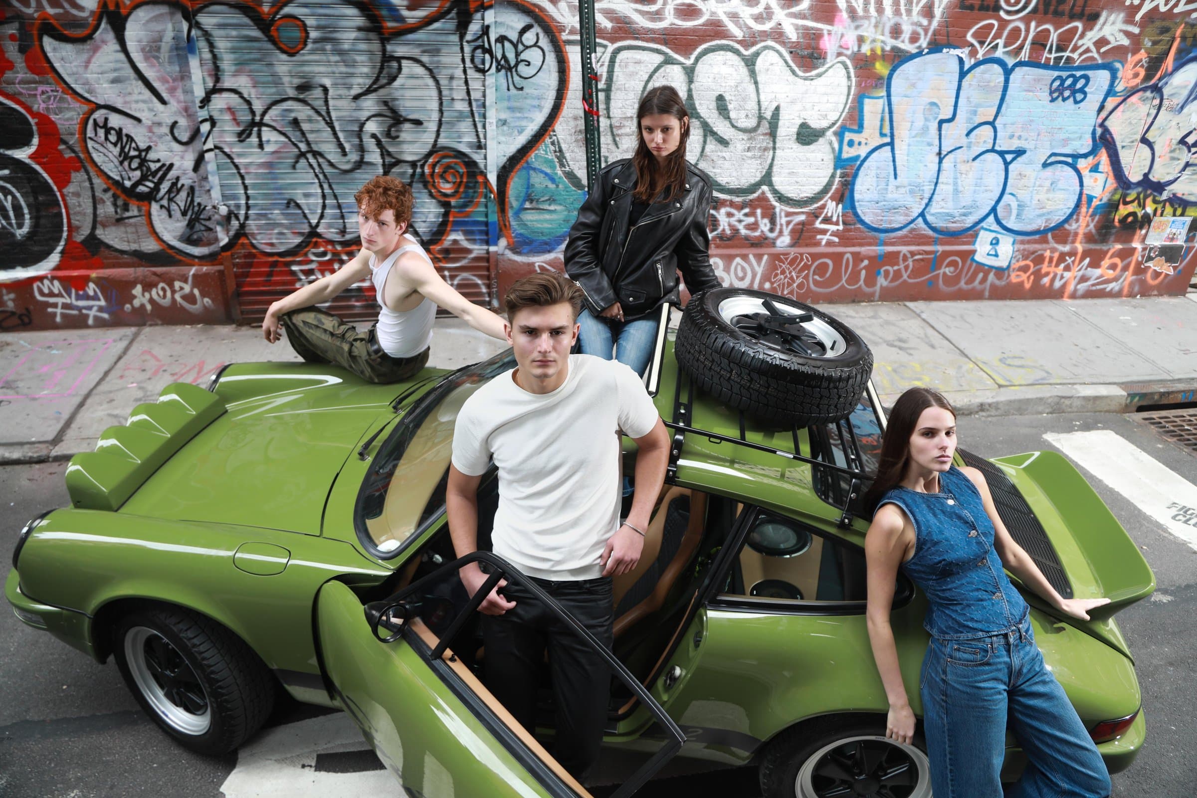 Four people around a green car beneath a graffiti wall